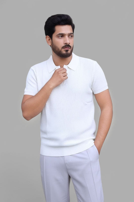 A man wearing a white short-sleeve ribbed knit polo shirt and light gray trousers, standing against a solid gray studio background.