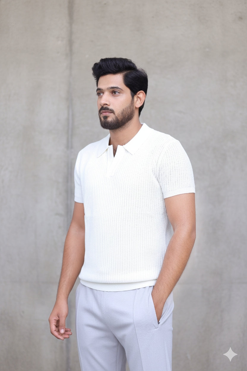 A man wearing a white ribbed short-sleeve polo shirt and light gray trousers, standing against a textured gray concrete wall.