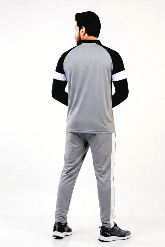 A man viewed from behind wearing a gray athletic tracksuit with black and white raglan sleeves and gray pants with white side stripes, standing against a plain white studio background.