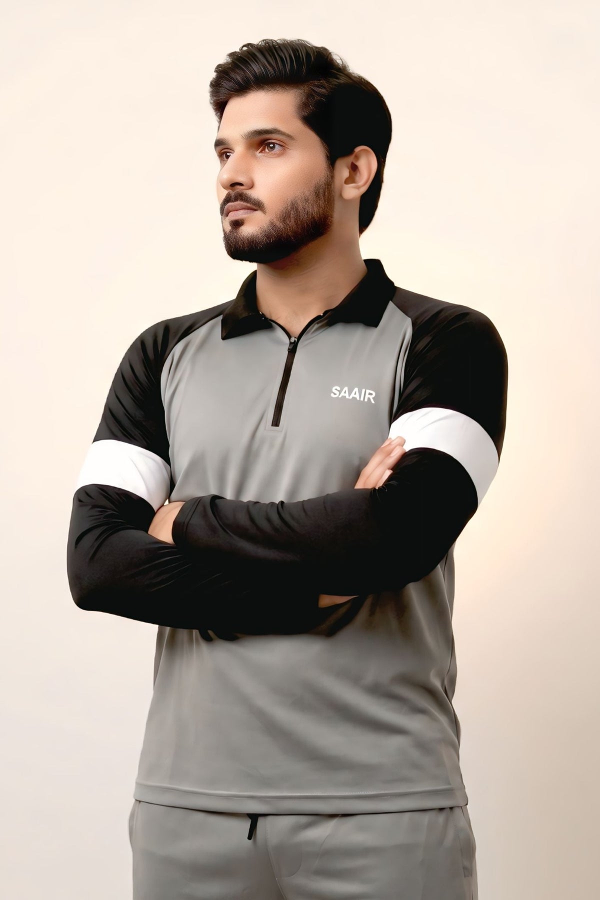 A man wearing a gray and black athletic long-sleeve shirt with white sleeve stripes and a small white logo on the chest, standing with arms crossed against a plain light background.