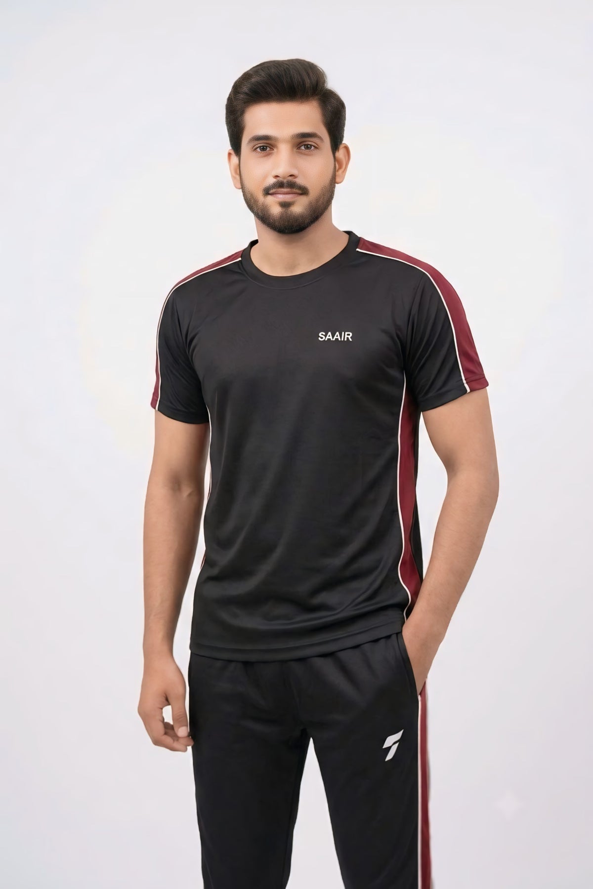 A man modeling a black short-sleeve athletic t-shirt with red side stripes and a white 'SAAIR' logo on the chest, paired with black track pants, against a light gray background.