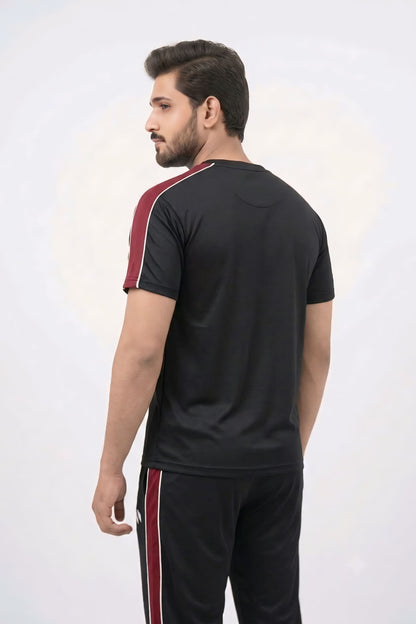 A man wearing a black athletic t-shirt and matching black track pants with red and white side stripes, shown from a three-quarters back view against a light gray studio background.