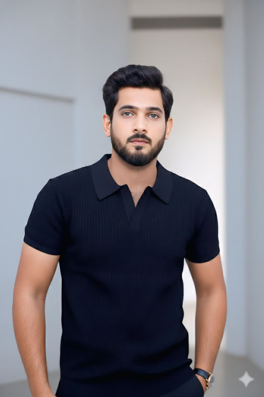 A man with a beard and styled hair wearing a dark navy ribbed short-sleeve polo shirt, standing against a softly blurred light gray background.
