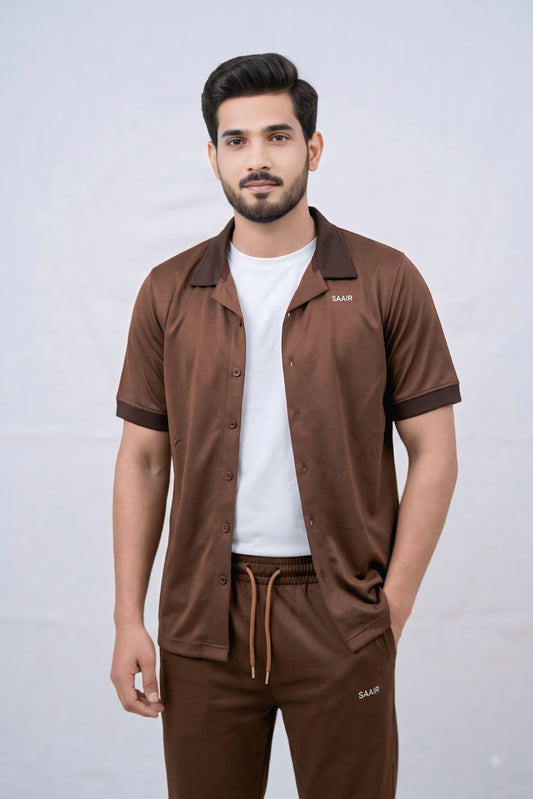 A man modeling a brown short-sleeve button-up shirt and matching brown drawstring pants set, worn over a white t-shirt, against a light gray studio background.