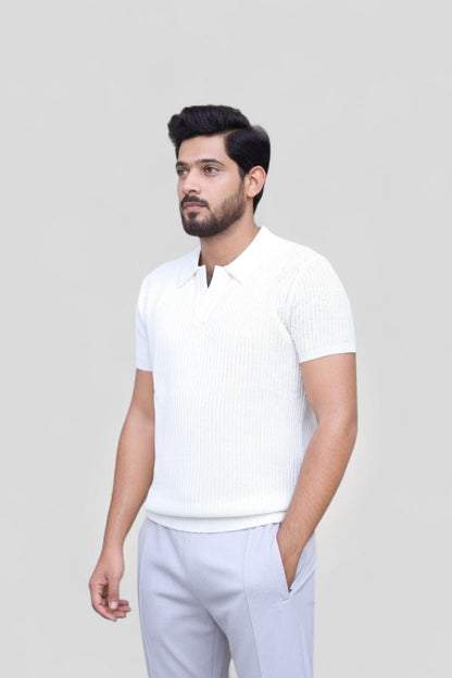 A man wearing a white short-sleeve ribbed knit polo shirt and light gray trousers, standing against a solid light gray background.