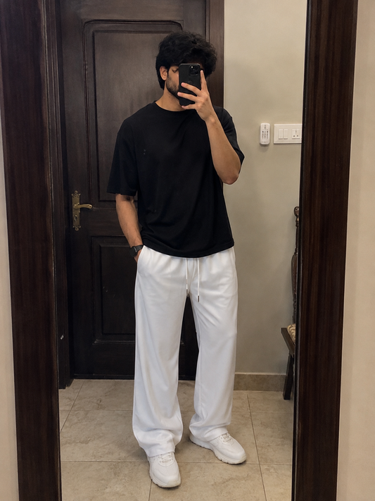 A man taking a mirror selfie wearing a black short-sleeve t-shirt and white drawstring sweatpants, standing in a doorway.
